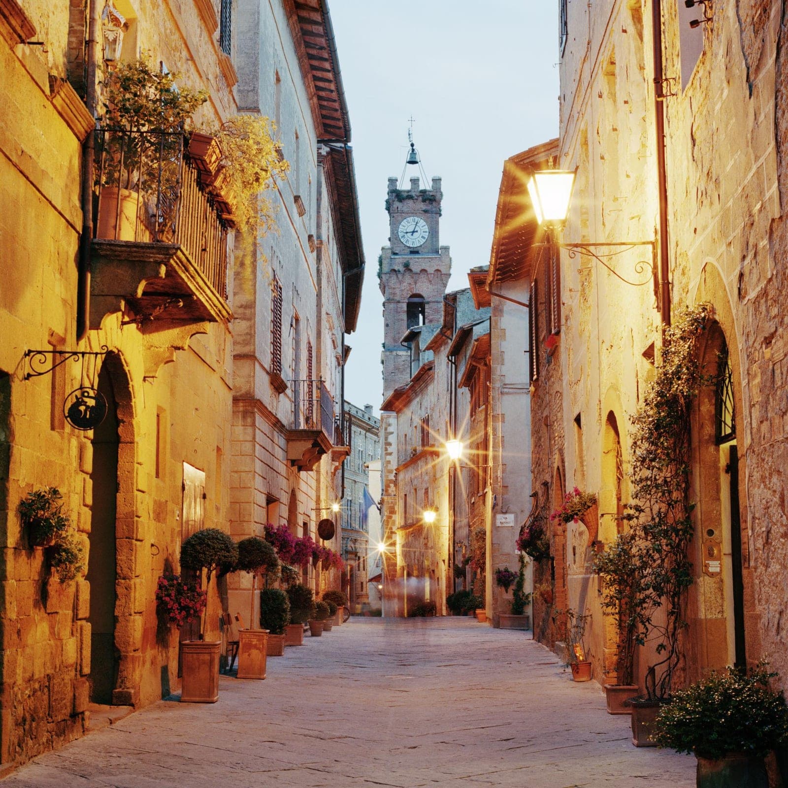 A narrow, lantern-lit stone street in Pienza leading toward the historic clock tower at dusk