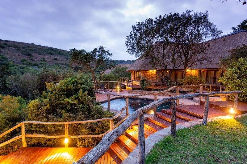A thatched-roof safari lodge with wooden walkways and a pool lit at dusk, surrounded by South African bushveld