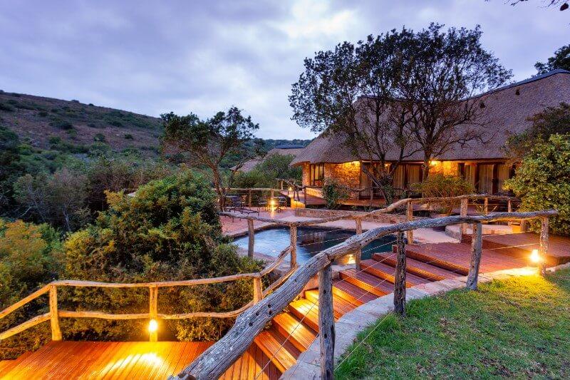 A thatched-roof safari lodge with wooden walkways and a pool lit at dusk, surrounded by South African bushveld