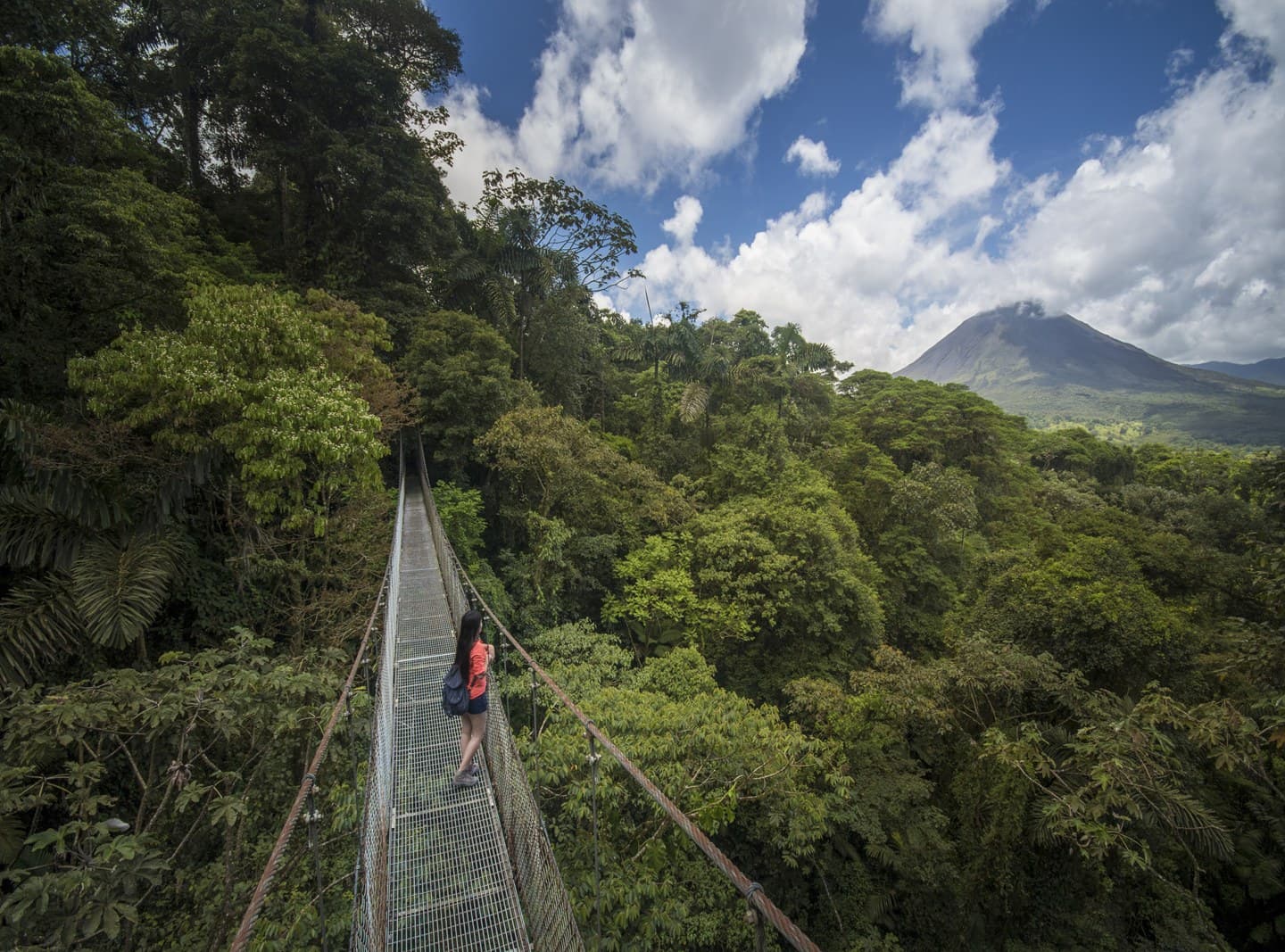 A hiker crossing a suspension bridge through the rainforest canopy toward Arenal Volcano
